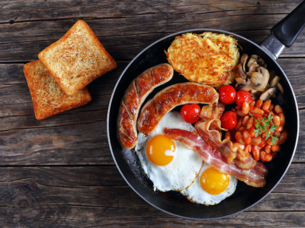 full english breakfast - bean, hash brown, fried eggs, bacon, roasted sausages, tomatoes, mushrooms on skillet on old dark wooden table with toasted bread, blank space left, view from above